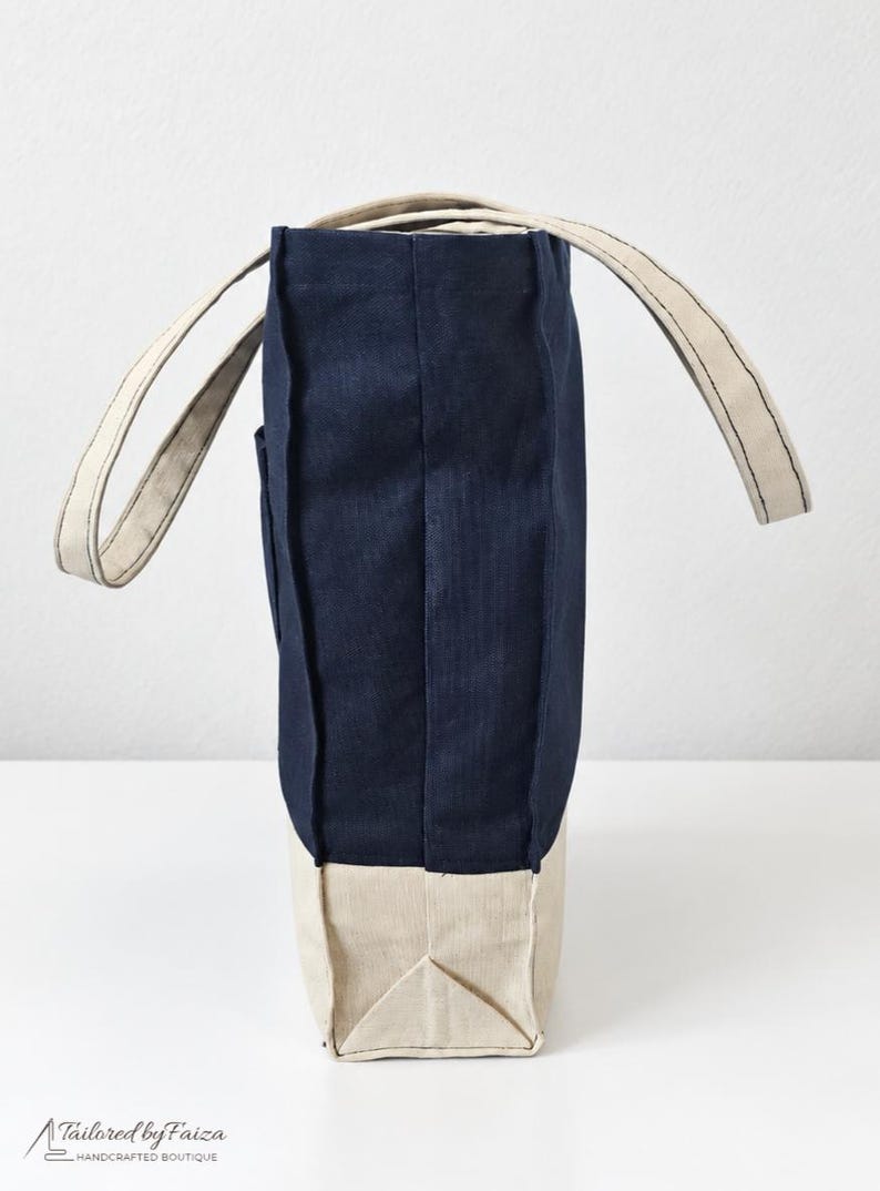 Puede incluir: Un bolso tote vertical con una secci&oacute;n superior azul marino profundo y una base inferior beige en contraste. El bolso incluye dos asas de tela beige y costuras visibles a lo largo de los lados. El texto Tailored by Faiza Handcrafted Boutique est&aacute; impreso en la esquina inferior izquierda.