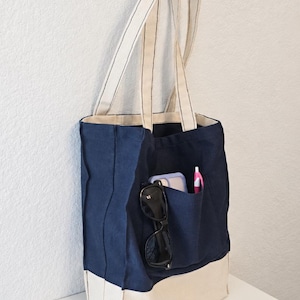 Puede incluir: Un bolso tote de lona azul marino con una base color crema y correas de hombro a juego. El exterior cuenta con un bolsillo frontal que contiene unas gafas de sol negras, un smartphone con funda morada clara y un bol&iacute;grafo rosa. Dise&ntilde;o estructurado.