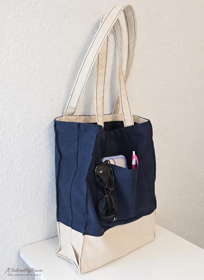 Puede incluir: Un bolso tote de lona azul marino con una base color crema y correas de hombro a juego. El exterior cuenta con un bolsillo frontal que contiene unas gafas de sol negras, un smartphone con funda morada clara y un bol&iacute;grafo rosa. Dise&ntilde;o estructurado.