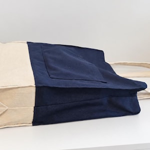 Puede incluir: Un bolso tote de lona bicolor con cuerpo principal azul marino, un bolsillo exterior grande y base y paneles laterales en color beige natural. El bolso incluye dos asas de tela beige a juego y muestra el texto Tailored by Faiza.