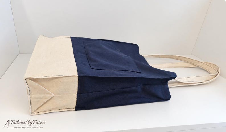 Puede incluir: Un bolso tote de lona bicolor con cuerpo principal azul marino, un bolsillo exterior grande y base y paneles laterales en color beige natural. El bolso incluye dos asas de tela beige a juego y muestra el texto Tailored by Faiza.