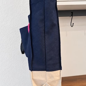 Puede incluir: Un bolso tote de lona azul marino con base y fuelles laterales en color beige natural. El bolso presenta un acento circular rosa brillante en el lateral e incluye asas de tela largas y claras. El texto Tailored by Faiza Handcrafted Boutique es visible.