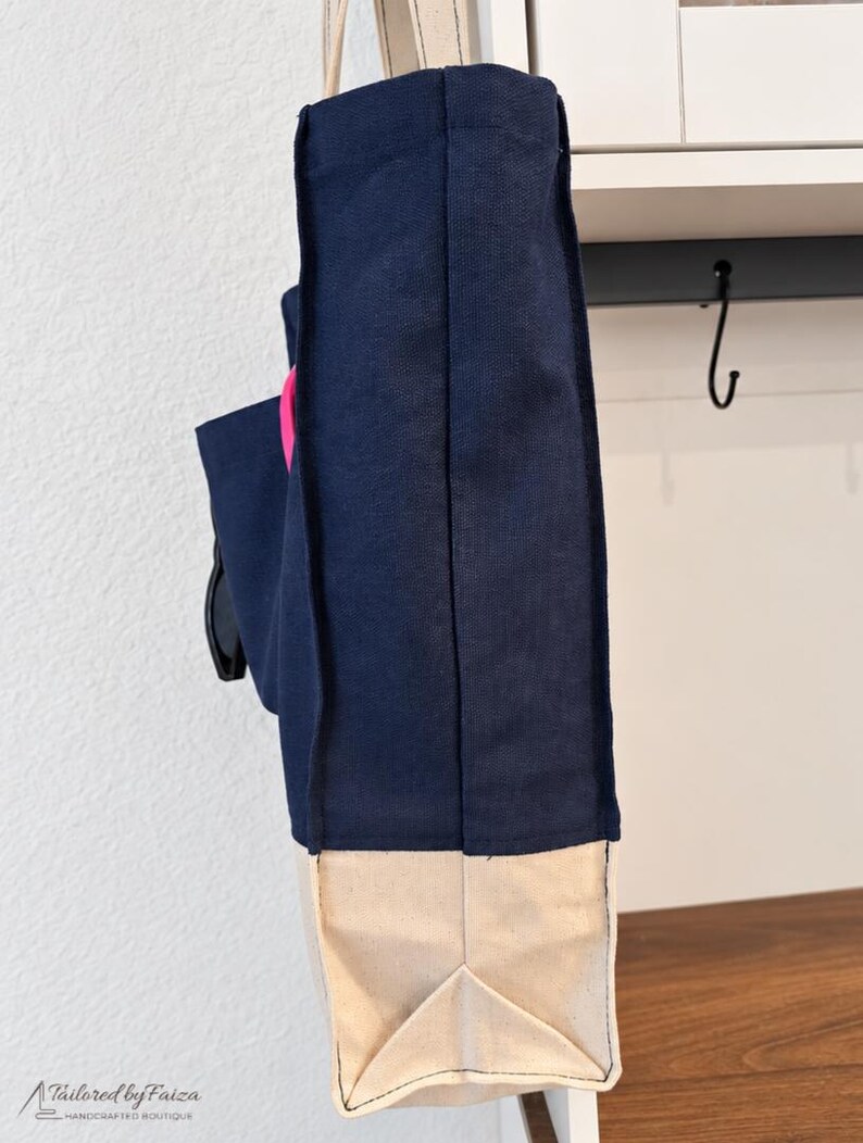 Puede incluir: Un bolso tote de lona azul marino con base y fuelles laterales en color beige natural. El bolso presenta un acento circular rosa brillante en el lateral e incluye asas de tela largas y claras. El texto Tailored by Faiza Handcrafted Boutique es visible.