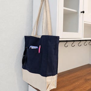 Puede incluir: Un bolso tote de lona en azul marino y crema con dos correas largas para el hombro. El bolso cuenta con un bolsillo exterior grande que contiene un smartphone morado claro, un bol&iacute;grafo rosa y unas gafas negras. El texto Tailored by Faiza es visible.