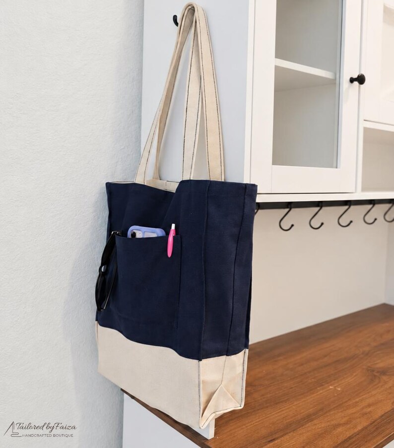 Puede incluir: Un bolso tote de lona en azul marino y crema con dos correas largas para el hombro. El bolso cuenta con un bolsillo exterior grande que contiene un smartphone morado claro, un bol&iacute;grafo rosa y unas gafas negras. El texto Tailored by Faiza es visible.