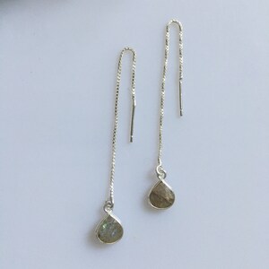 May include: A pair of silver threader earrings. Each earring has a fine chain that curves at the top and ends with a teardrop-shaped, iridescent gemstone. The gemstones display a mix of grey, green, and blue hues.