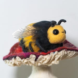 May include: A felt bumble bee with black and yellow stripes and clear wings sits on top of a red and white mushroom.