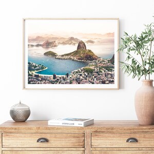 May include: An aerial view of Rio de Janeiro, Brazil, showing the iconic Sugarloaf Mountain and Guanabara Bay. The city is spread out below, with buildings and roads visible. The sky is a soft blue with a hint of pink.