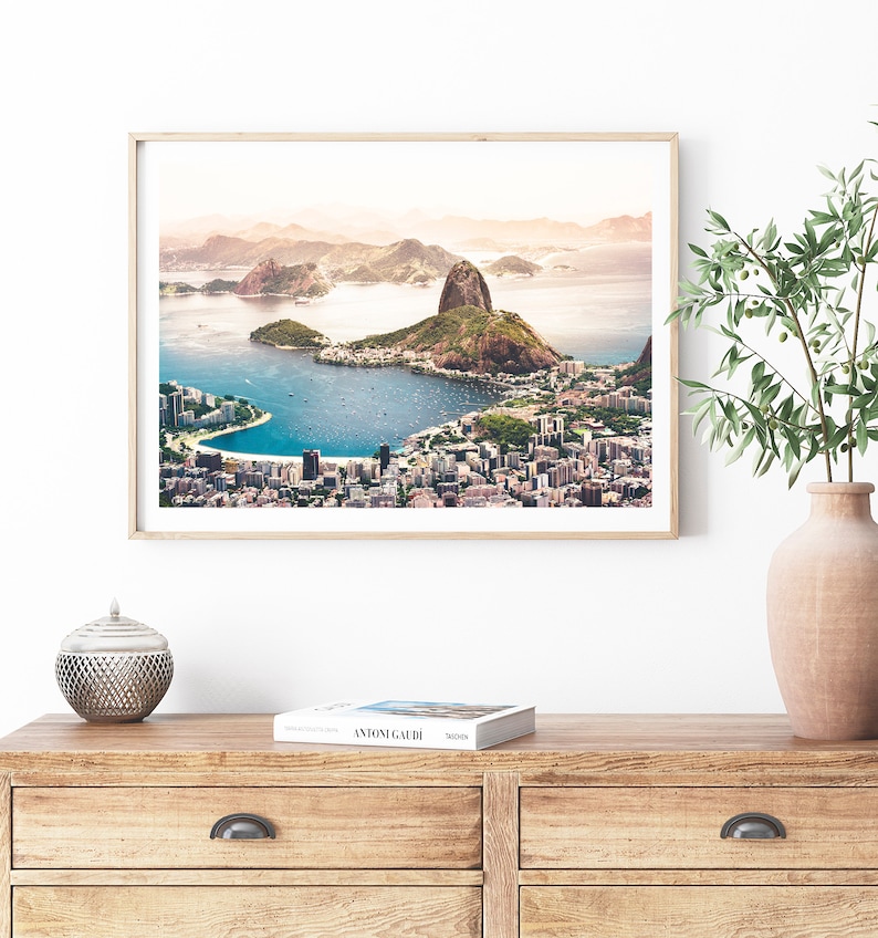 May include: An aerial view of Rio de Janeiro, Brazil, showing the iconic Sugarloaf Mountain and Guanabara Bay. The city is spread out below, with buildings and roads visible. The sky is a soft blue with a hint of pink.