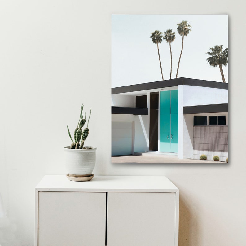 May include: A white mid-century modern house with a teal blue door. The house has a black roof and a black trim around the windows. There are palm trees in the background.