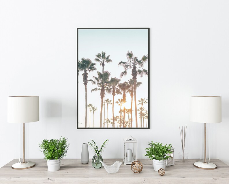 May include: A framed photograph of a row of palm trees against a light blue sky. The photo is displayed on a wooden shelf with two white lamps, two potted plants, a vase, a lantern, and a decorative bird sculpture.