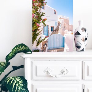 Puede incluir: Un edificio blanco con una puerta azul y flores rosas que crecen en el lateral. El edificio se encuentra en un lugar soleado con un cielo azul de fondo.