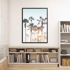 May include: A framed photograph of palm trees against a blue sky with a sunset in the background. The photograph is hanging on a white wall above a wooden bookshelf filled with books and decorative items.