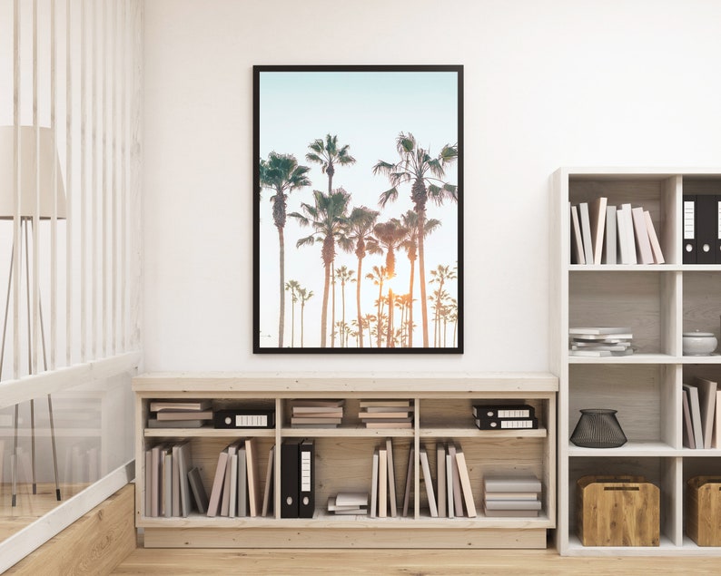 May include: A framed photograph of palm trees against a blue sky with a sunset in the background. The photograph is hanging on a white wall above a wooden bookshelf filled with books and decorative items.