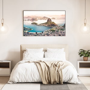 May include: A white bed with a beige throw blanket in a bedroom with a framed photograph of a city skyline above a body of water. The photograph is hanging on a white wall. There are two bedside tables with lamps on either side of the bed.