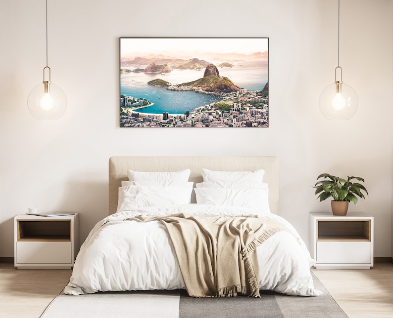 May include: A white bed with a beige throw blanket in a bedroom with a framed photograph of a city skyline above a body of water. The photograph is hanging on a white wall. There are two bedside tables with lamps on either side of the bed.