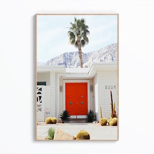 May include: A white mid-century modern house with a bright orange door and a palm tree in front. The house has a white brick exterior and a flat roof. The door has a circular design and is surrounded by a white frame. The palm tree is tall and slender, with green leaves. The house is set against a backdrop of mountains.