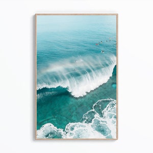 May include: An aerial view of a large wave breaking in turquoise ocean water. The wave is white and foamy, and there are several surfers in the water.