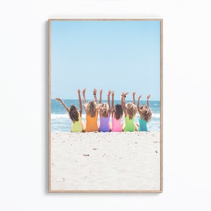 May include: Seven women in colorful swimsuits stand on a sandy beach with their arms raised in the air. The women are facing away from the camera and the ocean is in the background.
