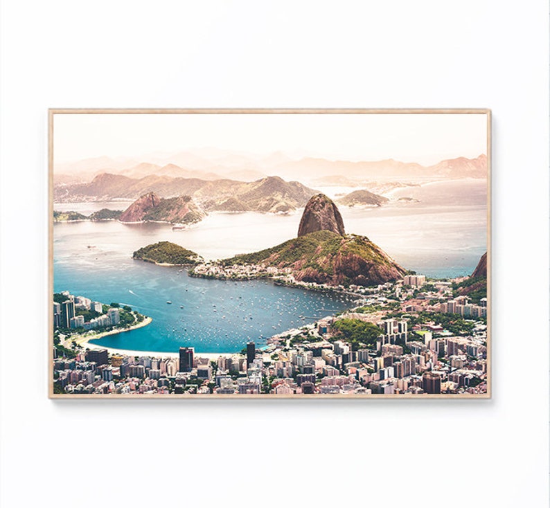 May include: An aerial view of a city with a large mountain in the background. The city is built on a peninsula with a bay of water in the foreground. The water is a light blue color and there are many boats in the bay. The mountain is a light brown color and is covered in vegetation. The sky is a light blue color with some clouds.