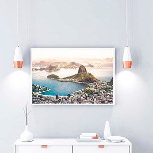 May include: A framed photograph of a city skyline with a large mountain in the background. The city is located on a peninsula with a body of water surrounding it. The photograph is in a white frame and is hanging on a white wall.
