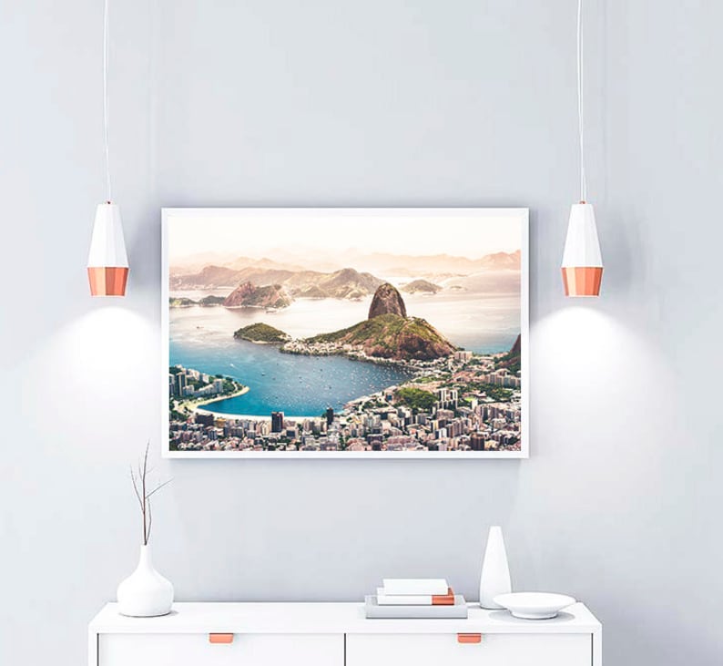 May include: A framed photograph of a city skyline with a large mountain in the background. The city is located on a peninsula with a body of water surrounding it. The photograph is in a white frame and is hanging on a white wall.