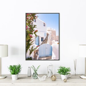 Puede incluir: Una fotograf&iacute;a enmarcada de un edificio blanco con una puerta azul y una enredadera trepadora con flores rosas. El edificio est&aacute; en un lugar soleado con un cielo azul.