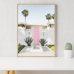 May include: A pink door with a white frame, surrounded by a white stucco house with palm trees in the background. The door is set in a white wall with a small fountain in front of it.
