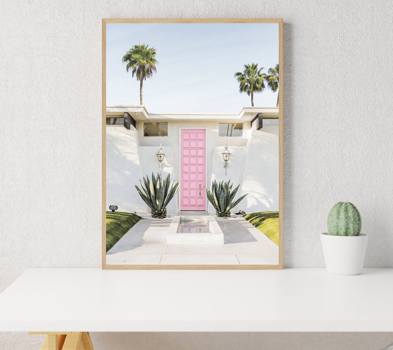 May include: A pink door with a white frame, surrounded by a white stucco house with palm trees in the background. The door is set in a white wall with a small fountain in front of it.