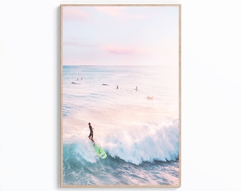 May include: A surfer rides a wave in the ocean at sunset. The water is a light blue and the sky is a soft pink and orange. There are other surfers in the distance.