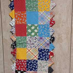 May include: A colorful, handmade quilt runner with a patchwork design. The runner features squares of fabric in various patterns, including polka dots, floral prints, and stripes, in shades of red, yellow, blue, and green. The edges are scalloped.