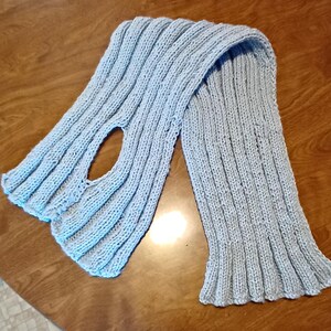 May include: A light blue knitted scarf with a hole in the center. The scarf is folded in half and laid flat on a wooden surface.