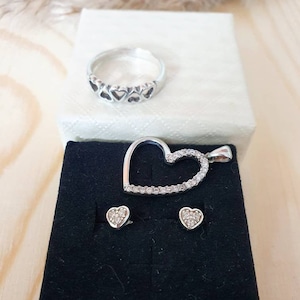 Op de afbeelding: Een set zilveren sieraden met een hartvormige hanger, een ring met een rij hartjes en een paar hartvormige oorbellen. Alle stukken zijn versierd met glinsterende, heldere stenen.