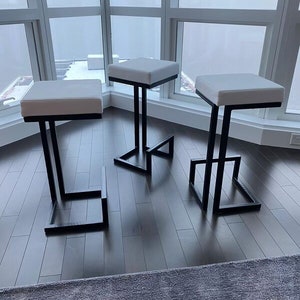May include: Three white cushioned bar stools with black metal frames. The stools are arranged in a row on a dark wood floor.