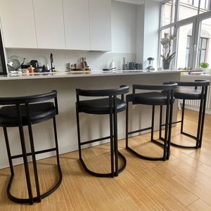 Op de afbeelding: Vier zwarte metalen barstoelen met zwarte leren zittingen. De stoelen staan in een rij in een keuken.