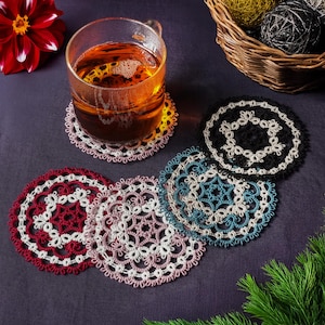 Puede incluir: Cinco manteles individuales de ganchillo en diferentes colores, incluyendo rojo, negro, blanco y azul, están dispuestos alrededor de un vaso de té sobre una superficie de color morado oscuro.