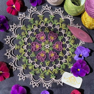 May include: A delicate white lace doily with a floral design in shades of green, purple, and yellow. The doily is surrounded by purple pansies and a tag that reads "AYA Lace".