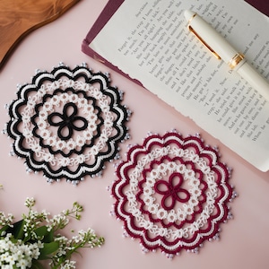 Pode incluir: Duas bases de copo circulares feitas à mão. Uma é preta e branca, a outra bordô e branca. Ambas têm um centro floral e bordas com contas. Elas são colocadas em uma superfície rosa com um livro e uma caneta.