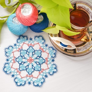 May include: A handmade, intricate doily in shades of blue and coral, with a floral design. Three spools of thread in matching colors are nearby, along with a tatting shuttle and a cup of tea. Green leaves add a natural touch to the scene.