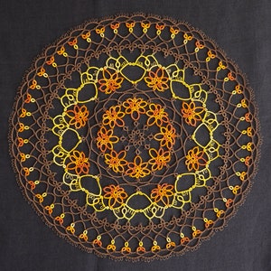 May include: A detailed, circular doily with a complex lace pattern. The doily features concentric rings of brown, yellow, and orange thread, creating a floral design. The intricate lacework is set against a dark background.