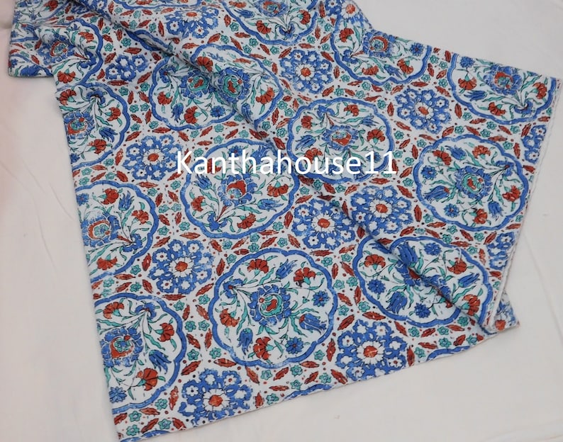 May include: White fabric with a blue and red floral pattern. The fabric has a repeating pattern of stylised flowers and leaves. The fabric is folded in half and the edges are visible.