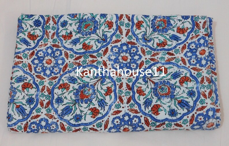 May include: White cotton fabric with a blue and red floral pattern. The fabric has a distressed look and features a repeating pattern of stylised flowers and leaves. The text "Kantha house 11" is visible in the centre of the fabric.