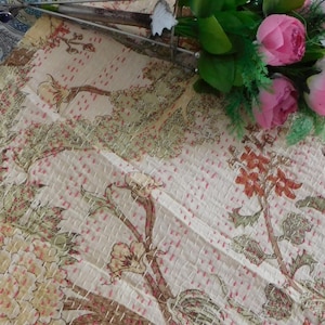 Puede incluir: Una colcha kantha de color crema con un patr&oacute;n floral en tonos marrones, verdes y rosas. La colcha est&aacute; cosida a mano con hilo rosa y tiene una superficie texturizada. La colcha se muestra en una mesa con un ramo de flores rosas.