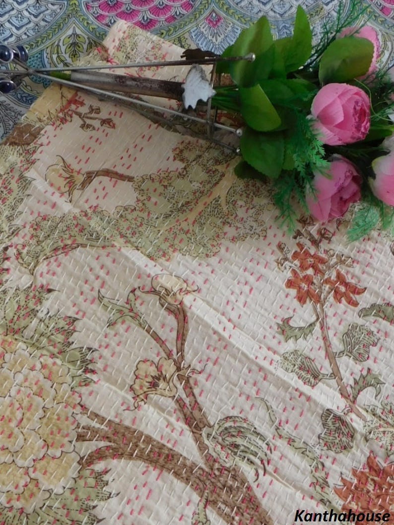 Puede incluir: Una colcha kantha de color crema con un patr&oacute;n floral en tonos marrones, verdes y rosas. La colcha est&aacute; cosida a mano con hilo rosa y tiene una superficie texturizada. La colcha se muestra en una mesa con un ramo de flores rosas.