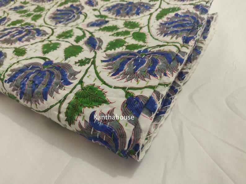 May include: White cotton fabric with a blue and green floral print. The fabric is folded and has a stitched edge. The text "Kantha house" is visible on the fabric.