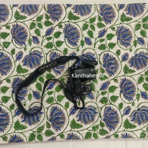May include: White cotton kantha quilt with a blue and green floral pattern. The quilt is hand-stitched and has a traditional Indian design. The quilt is shown with a camera and strap on top of it.