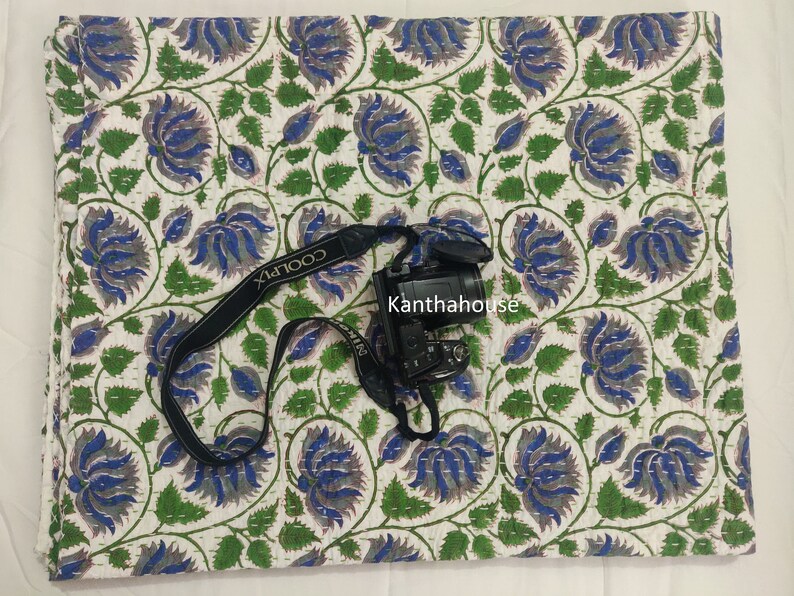 May include: White cotton kantha quilt with a blue and green floral pattern. The quilt is hand-stitched and has a traditional Indian design. The quilt is shown with a camera and strap on top of it.