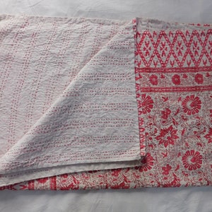 Op de afbeelding: Een wit kantha-deken met rode bloemen- en geometrische patronen. Het deken is dubbelgevouwen, waardoor de stiksels aan de witte kant zichtbaar zijn.