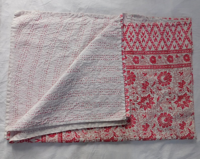 Op de afbeelding: Een wit kantha-deken met rode bloemen- en geometrische patronen. Het deken is dubbelgevouwen, waardoor de stiksels aan de witte kant zichtbaar zijn.