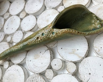 May include: A green ceramic spoon with a textured surface and a speckled glaze. The spoon is shaped like a seashell and has a wide, shallow bowl.